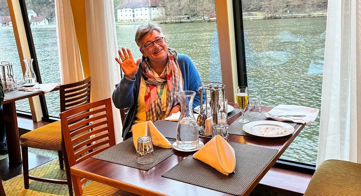 Reiseteilnehmerin im Restaurant des A-ROSA Flusskreuzfahrtschiffs winkt fröhlich in die Kamera