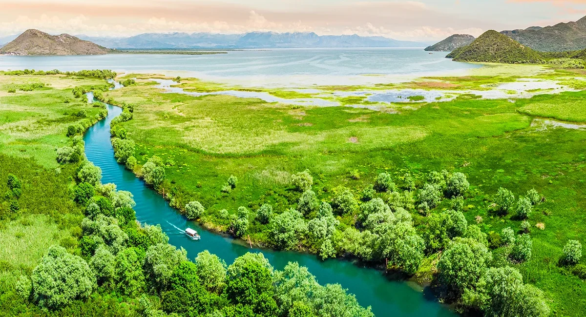 Der Skadar-Nationalpark in Albanien mit Blick auf den Skadarsee und seiner malerischen Bergkette im Hintergrund. Ein Fluss, auf dem ein kleines Fischerboot fährt, schlängelt sich durch die grüne Landschaft.