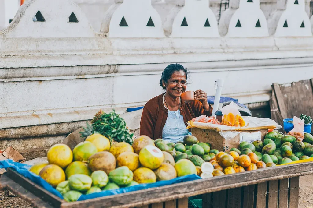 Eine Marktfrau aus Sri Lanka verkauft Früchte an ihrem Stand und lächelt in die Kamera.