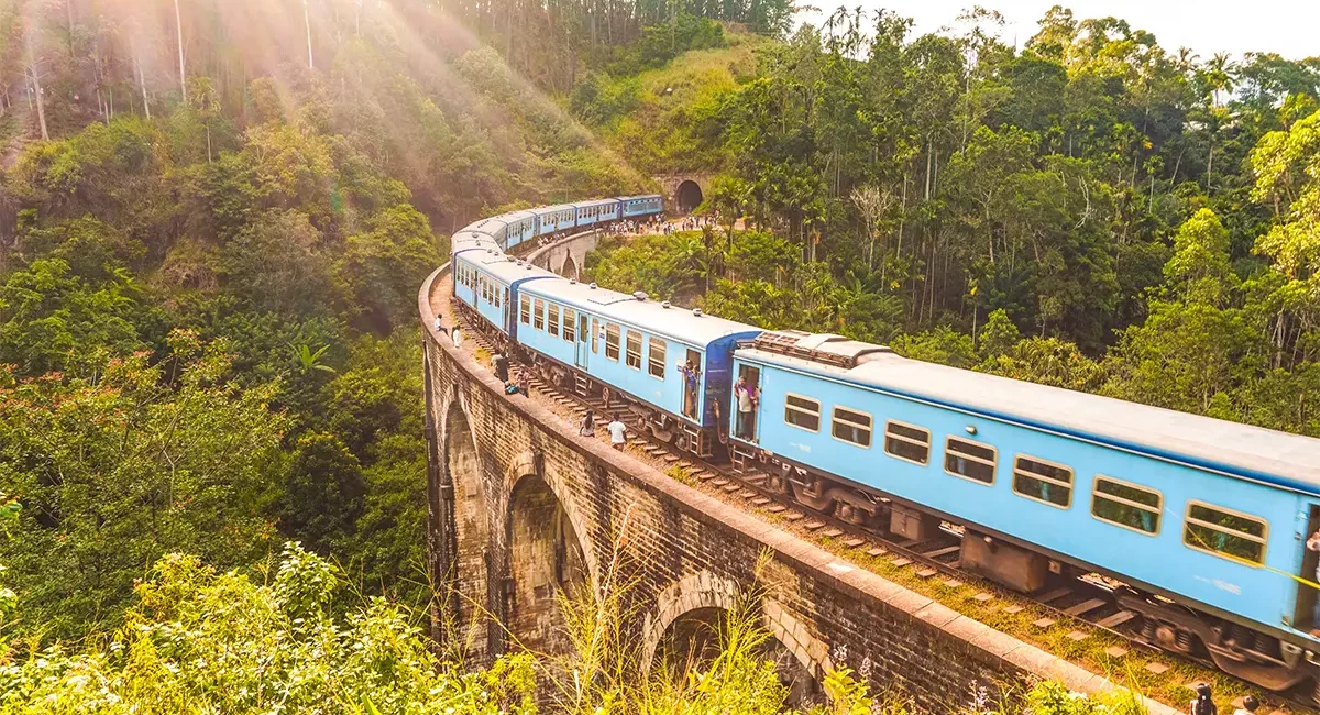 Ein blauer Zug fährt über die historische Nine Arch Bridge inmitten der üppigen, grünen Hügellandschaft von Sri Lanka.