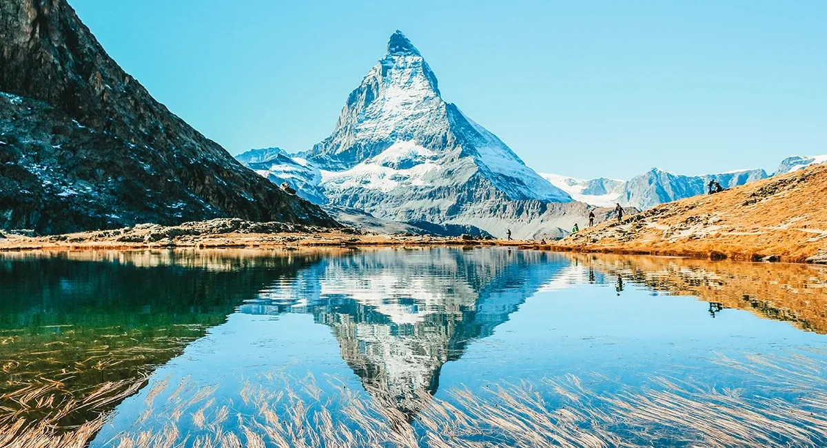 Das schneebedeckte Matterhorn, das sich perfekt im klaren Wasser des Riffelsees spiegelt, während sich einige Wanderer am Uferbereich aufhalten.