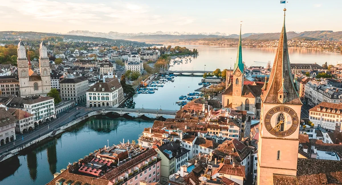Die Hauptstadt Zürich liegt in warmem Licht, mit ihren markanten Türmen und dem historischen Zentrum. Dahinter erstrecken sich der Zürichsee und eine schneebedeckte Bergkette.