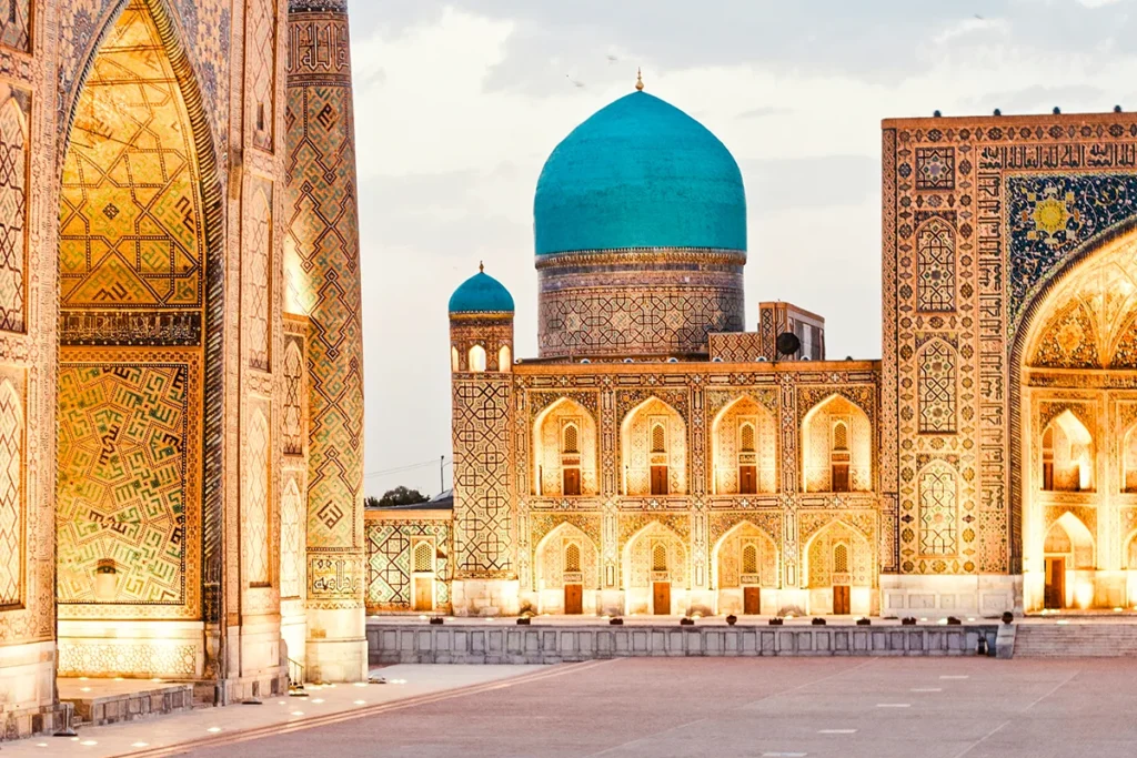 Historisches usbekisches Gebäude in Samarkand mit türkisblauer Kuppel und kunstvollen Fliesen an den Fassaden, die hell beleuchtet sind.