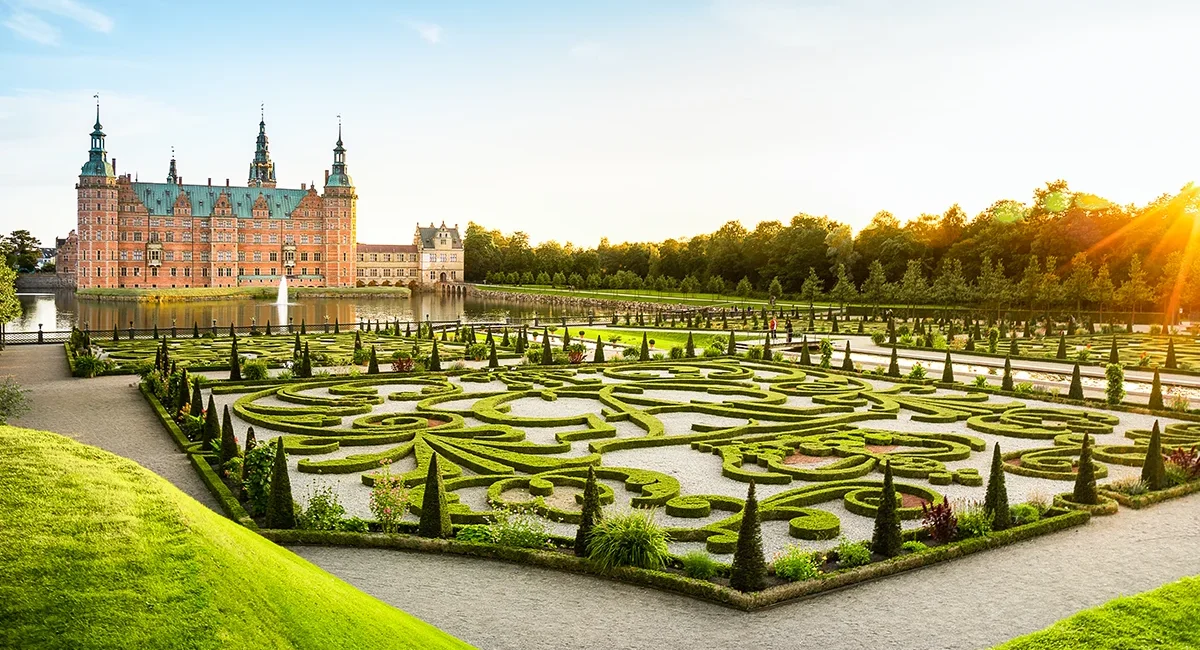 Das große, rote Backsteinschloss Frederiksborg in Dänemarkt, umgeben von einem kunstvoll angelegten formalen Barockgarten im Vordergrund.