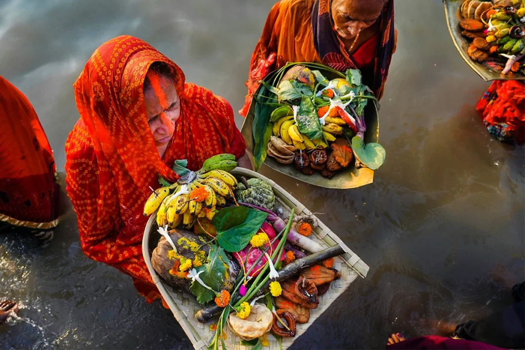 Frauen in traditioneller Kleidung stehen im Wasser eines Flusses in Indien und halten Körbe mit Opfergaben, was auf eine religiöse Zeremonie hindeutet.