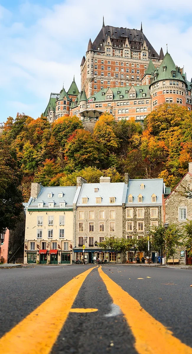Eine Straße führt durch die Unterstadt auf ein großes, schlossähnliches Gebäude zu, welches auf einem mit herbstlichen Bäumen bewachsenen Hügel in Québec-Stadt, Kanada thront.