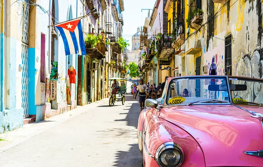 Eine enge Straße in Havanna, Kuba, mit einem rosafarbenen Oldtimer im Vordergrund und einer kubanischen Flagge, die über farbenfrohen Gebäuden weht.