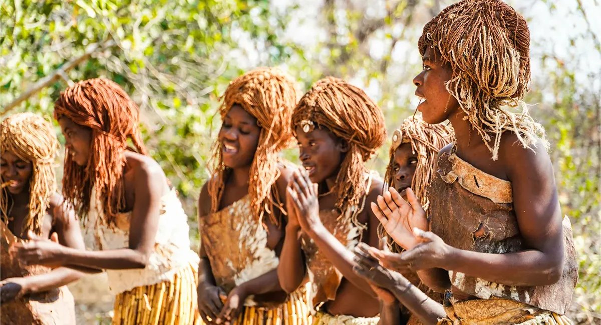 Eine Gruppe von einheimischen Frauen aus Namibia, bekleidet mit traditionellen Röcken aus Schilf und Leder, führt im Freien einen kulturellen Tanz auf und klatscht dazu rhythmisch in die Hände.