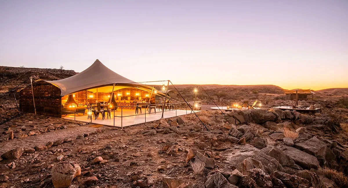 Ein Wüstencamp in der trockenen, felsigen Landschaft Namibias, mit einem offenen, stimmungsvoll beleuchteten Pavillon im warmen Licht des Sonnenuntergangs.