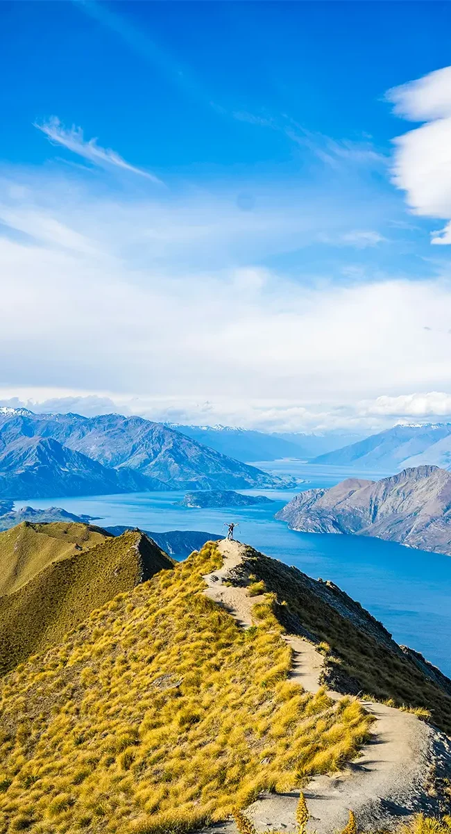 Ein Wanderer auf dem Kamm des Roys Peak in Neuseeland, der den atemberaubenden Panoramablick auf den darunter liegenden Lake Wānaka und die umliegenden Berge genießen.