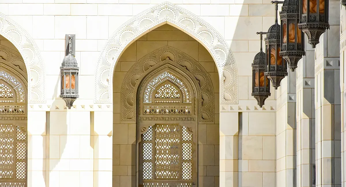 Architektonische Details einer Moschee im Oman, mit ihren charakteristischen weißen Marmorwänden, kunstvollen Steinmetzarbeiten an den Bögen und Gittern sowie den hängenden Bronzelaternen.