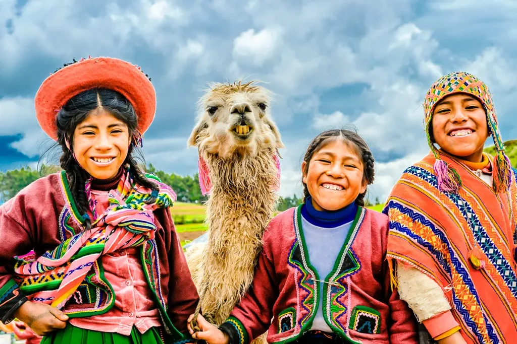Drei lachende Kinder in traditioneller Andenkleidung zusammen mit einem Alpaka vor einer Landschaftskulisse in Peru.