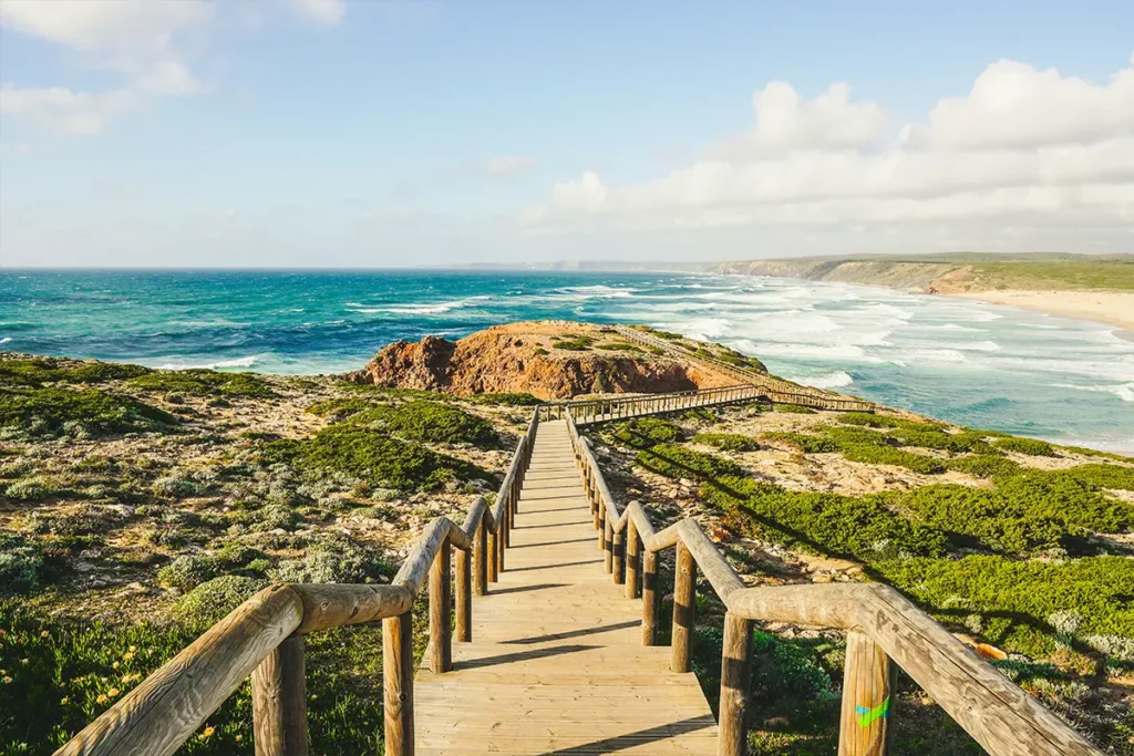 Ein langer Holzsteg, der über eine bewachsene Klippenlandschaft zu einem weitläufigen Sandstrand und dem Atlantik in Portugal führt.