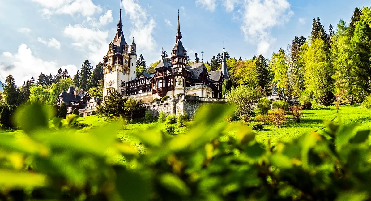 Das rumänische Schloss Peleș steht mit seinen Türmen und dem Fachwerkstil in einer grünen Landschaft mit vielen Bäumen.