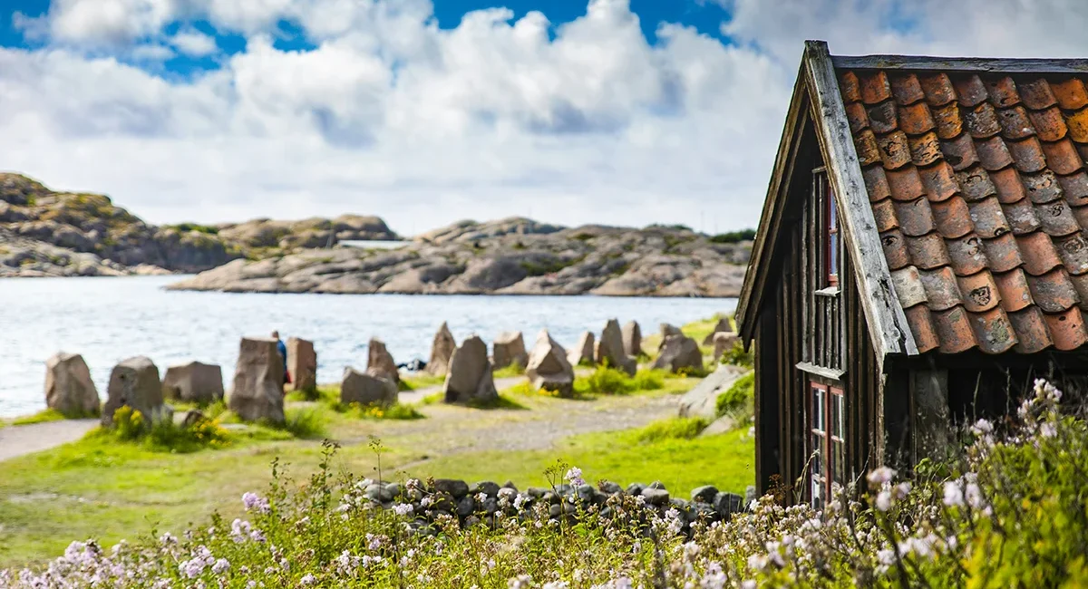 Eine alte kleine Holzhütte steht am Ufer eines Sees in Schweden, umgeben von Schärenlandschaften und Granitfelsen.