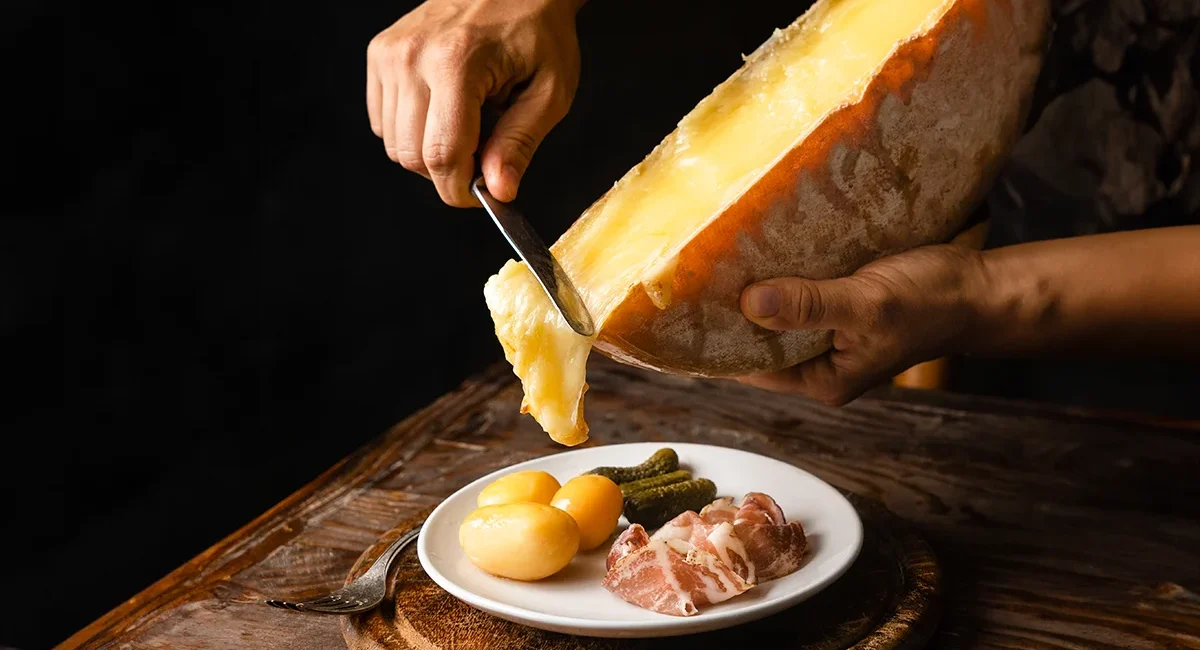 Mann schabt mit einem Messer schweizer Raclette-Käse auf Kartoffeln und Speck, die auf einem Teller liegen
