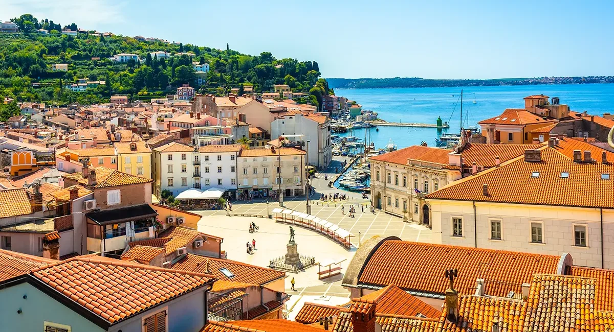 Die slowenische Küstenstadt Piran mit ihren dicht gedrängten Gebäuden mit orangefarbenen Ziegeldächern und dem Hafen.