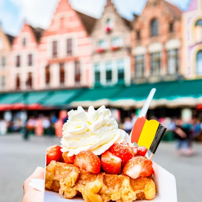 Auf einem Marktplatz mit bunten Häusern wird eine belgische Waffel mit Erdbeeren und Sahne gehalten.