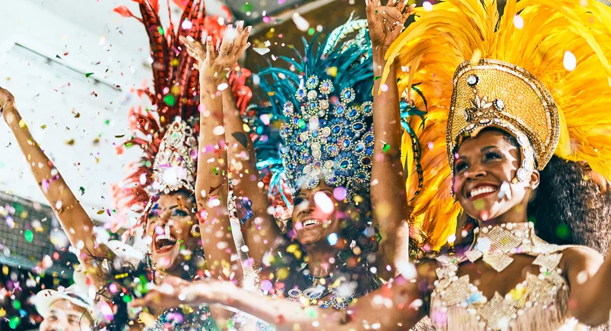 Drei bunt kostümierte Samba-Tänzerinnen beim brasilianischen Karneval mit viel Konfetti im Vordergrund.