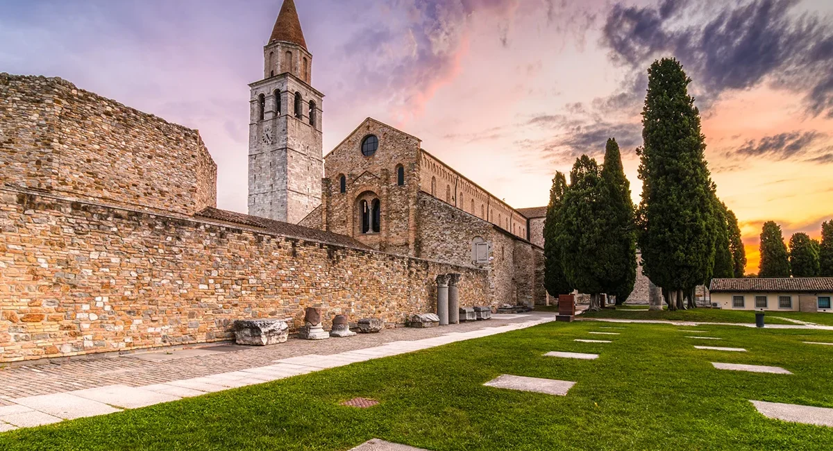 Die Basilika Santa Maria Assunta in Aquileia in Italien mit ihrem markanten Glockenturm und einer angrenzenden Grünanlage während eines stimmungsvollen Sonnenuntergangs.