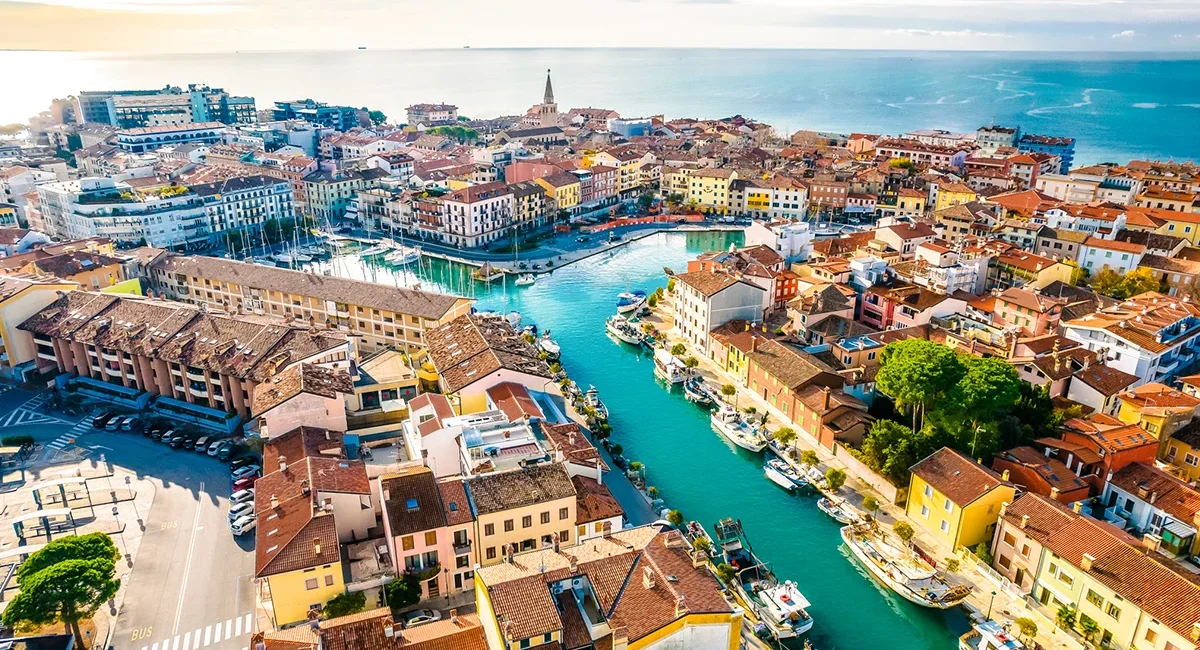 Die italienischen Stadt Grado, auf der ein von Booten gesäumter Kanalhafen zu sehen ist, der durch ein dicht bebautes Stadtzentrum mit historischen Gebäuden führt und im Hintergrund an die Adria grenzt.