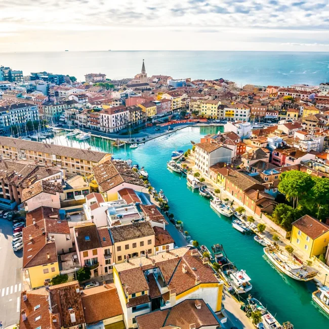 Die italienischen Stadt Grado, auf der ein von Booten gesäumter Kanalhafen zu sehen ist, der durch ein dicht bebautes Stadtzentrum mit historischen Gebäuden führt und im Hintergrund an die Adria grenzt.