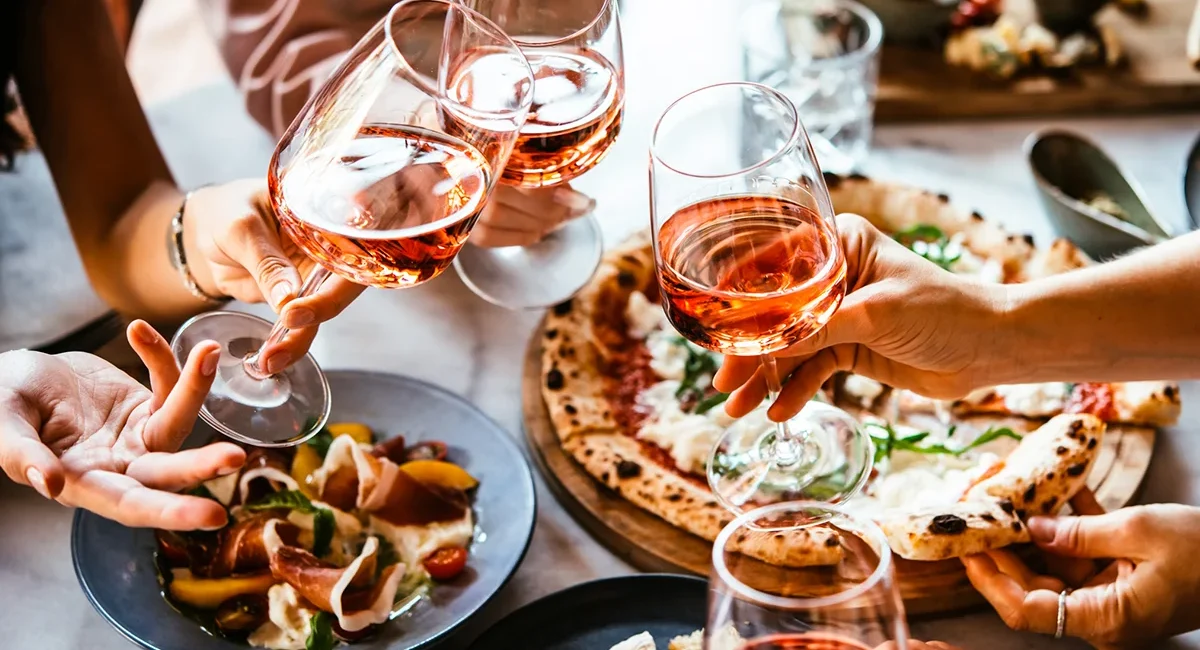 Eine Freundesgruppe sitzt am Tisch eines italienischen Restaurants, isst Pizza und stößt mit vier Gläsern Roséwein an.