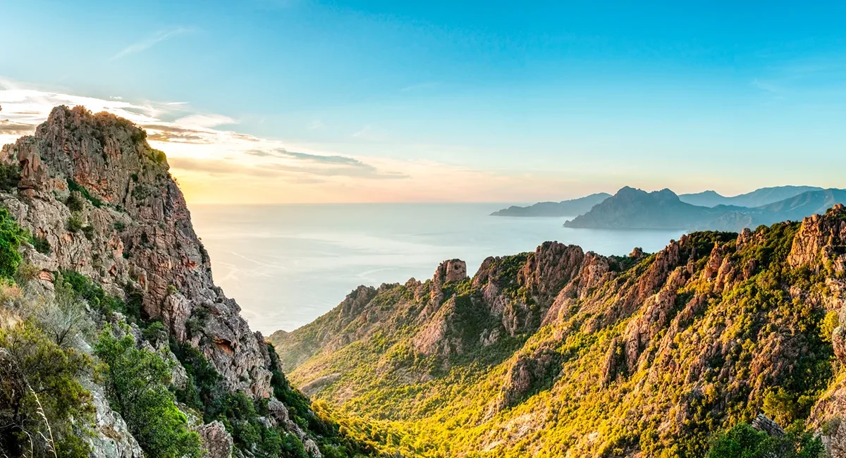 Die zerklüftete, rötliche Felsenlandschaft der Calanques de Piana auf Korsika, die bei tiefstehender Sonne über dem glitzernden Mittelmeer thront.