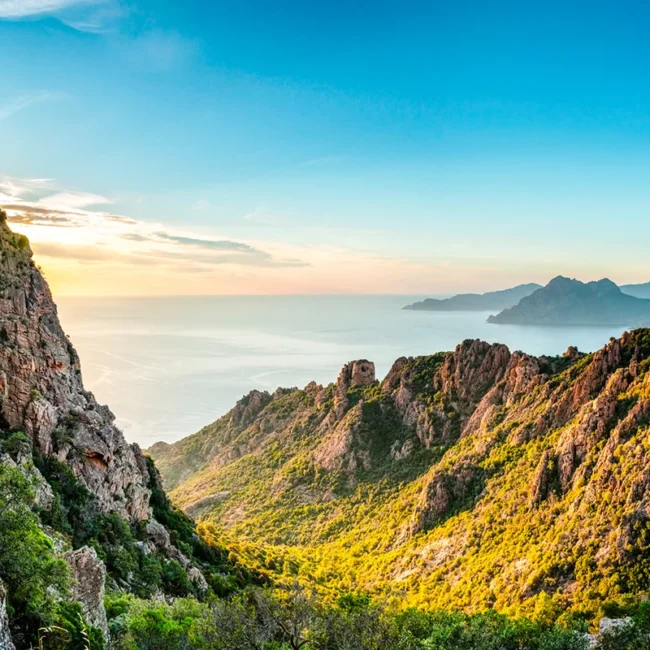Die zerklüftete, rötliche Felsenlandschaft der Calanques de Piana auf Korsika, die bei tiefstehender Sonne über dem glitzernden Mittelmeer thront.