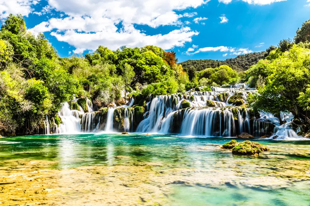Der Wasserfall Skradinski buk im kroatischen Nationalpark Krka, dessen klares, türkisfarbenes Wasser über mehrere Travertinstufen in ein von dichter, grüner Vegetation umgebenes Becken stürzt.