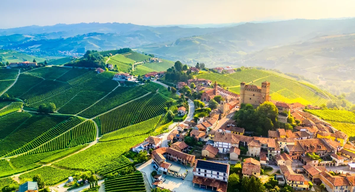 Das italienische Dorf Serralunga d'Alba im Piemont mit seiner markanten Burg auf einem Hügel, umgeben von weiten, grünen Weinbergen im Sonnenlicht.