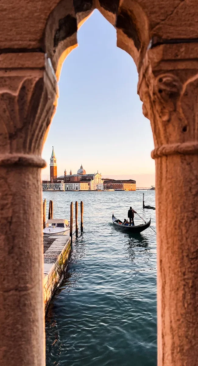 Ein verzierter venezianischer Steinbogen in Venedig mit einer Gondel im Wasser und der Insel San Giorgio Maggiore im Hintergrund.
