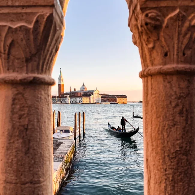 Ein verzierter venezianischer Steinbogen in Venedig mit einer Gondel im Wasser und der Insel San Giorgio Maggiore im Hintergrund.