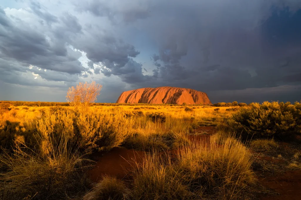Der Ayers Rock (Uluru) leuchtet in warmem, rötlichem Licht, während darüber ein dunkler, wolkiger Himmel hängt. Das stimmungsvolle Licht hebt den Felsen eindrucksvoll aus der weiten Landschaft hervor.