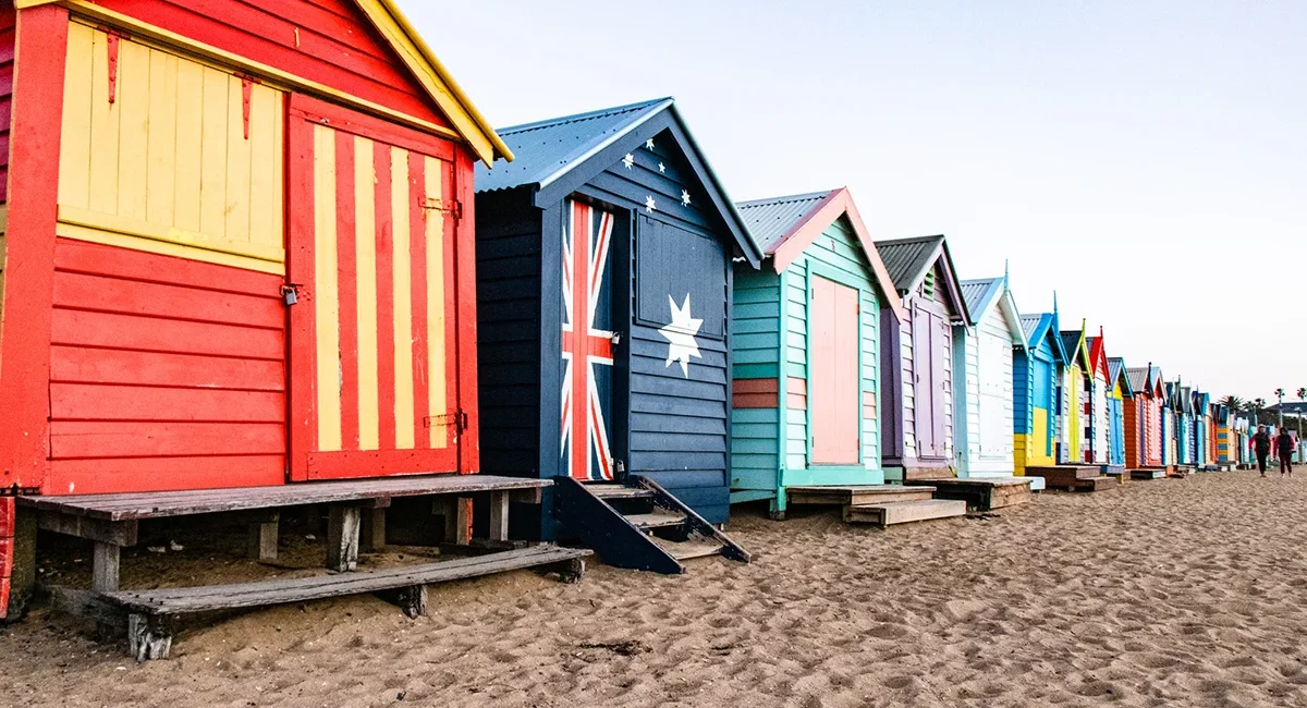 „Bunte Strandhütten stehen in einer Reihe am Brighton Beach in Melbourne. Die kleinen Holzhäuschen sind in verschiedenen Farben gestrichen und liegen direkt am Sandstrand vor dem Meer