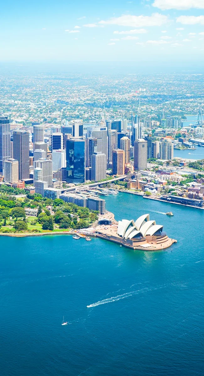 Luftaufnahme von Sydney bei Tageslicht: Das Opernhaus mit seinen weißen, segelförmigen Dächern liegt am Hafen neben der Harbour Bridge. Umgeben von blauem Wasser, Booten und einer dichten Skyline moderner Hochhäuser