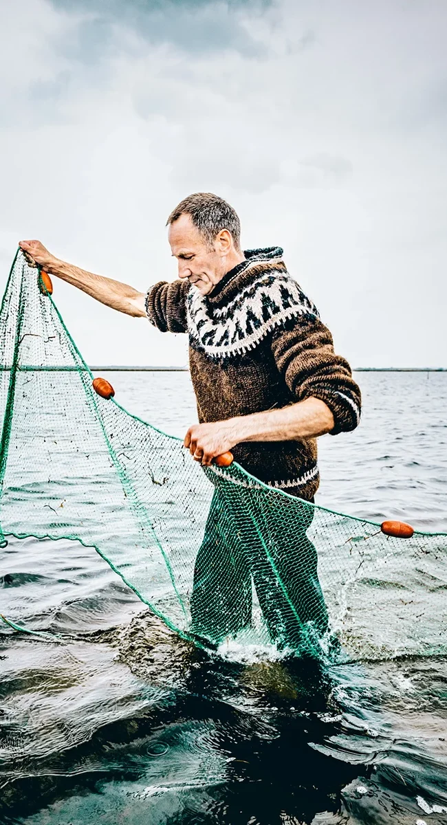 Ein Fischer steht im flachen Wasser des Meeres vor der Küste Dänemarks und ist mit seinem Fanggerät beschäftigt. Um ihn herum erstreckt sich das offene Meer.