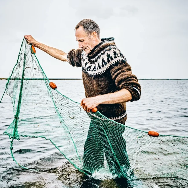 Ein Fischer steht im flachen Wasser des Meeres vor der Küste Dänemarks und ist mit seinem Fanggerät beschäftigt. Um ihn herum erstreckt sich das offene Meer.