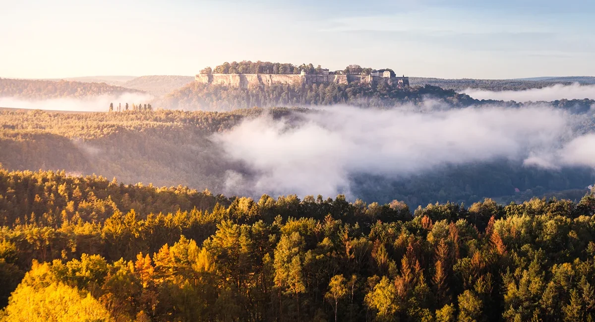 Die über einem morgendlichen Nebelmeer thronende Festung Königstein inmitten der herbstlichen Waldlandschaft des Elbsandsteingebirges.