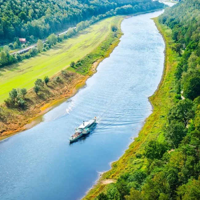 Ein Ausflugsschiff fährt an einem sonnigen Tag entlang der Elbe und vorbei an grünen Ufern.