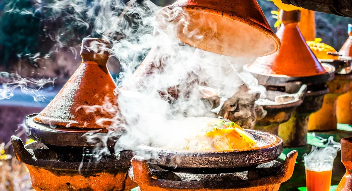 Eine traditionelle marokkanische Tajine steht in einem Tongefäß auf dem Tisch. Aus dem Gericht steigt heißer Dampf auf.
