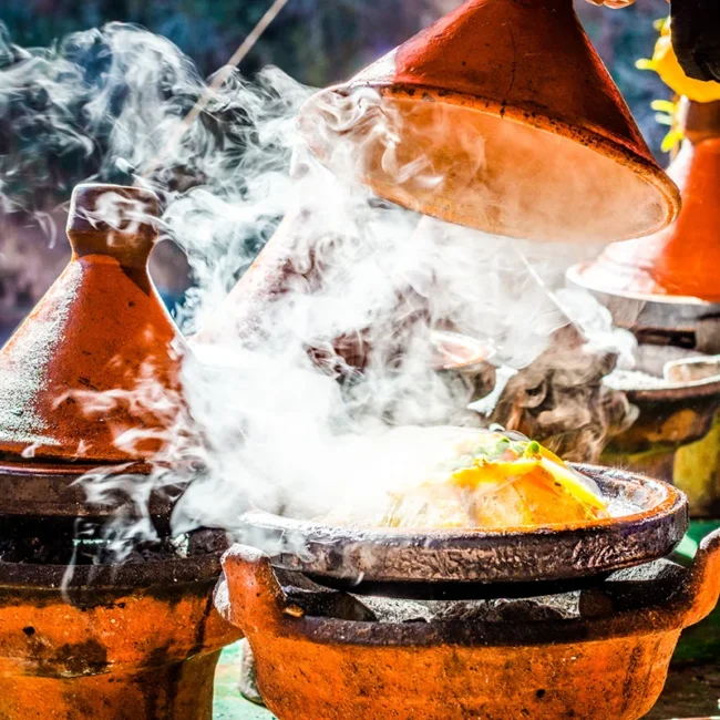 Eine traditionelle marokkanische Tajine steht in einem Tongefäß auf dem Tisch. Aus dem Gericht steigt heißer Dampf auf.