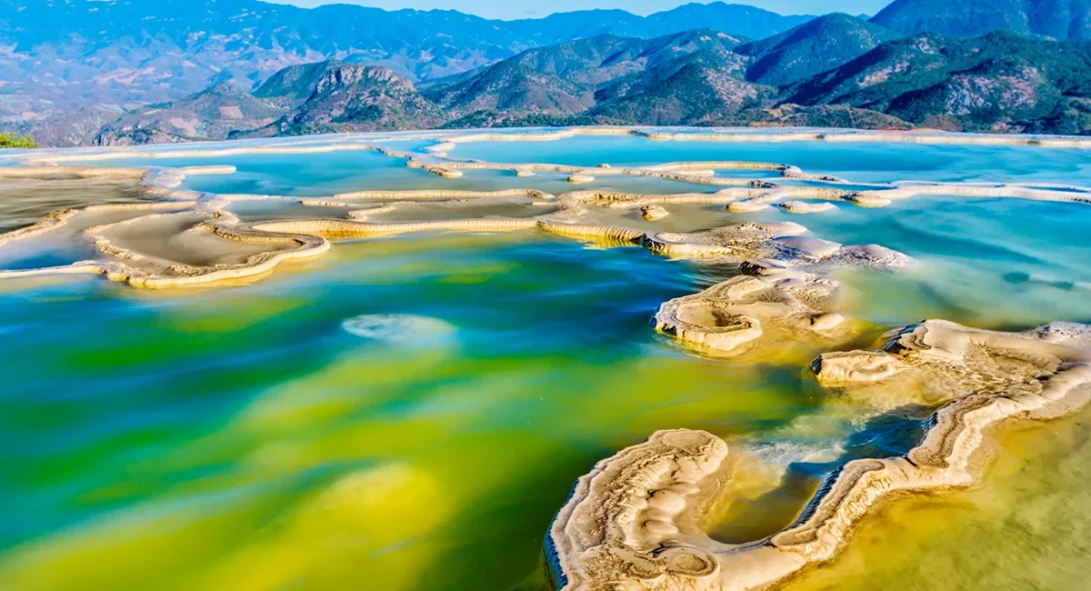 Blick von oben auf Hierve el Agua in Oaxaca, Mexiko. Die natürlichen türkisfarbenen Wasserbecken liegen auf einer Felsformation über der umliegenden Landschaft.