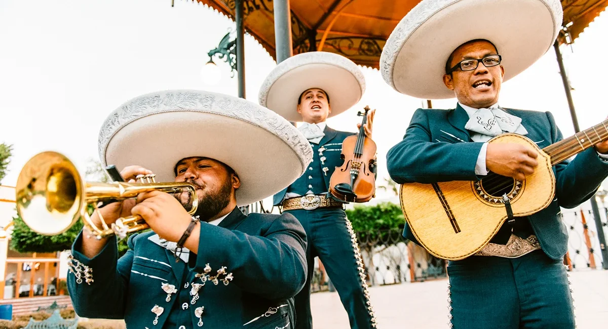 Drei Mariachi-Musiker tragen große weiße Sombreros und traditionelle Kleidung. Sie spielen Trompete, Geige und Gitarre.