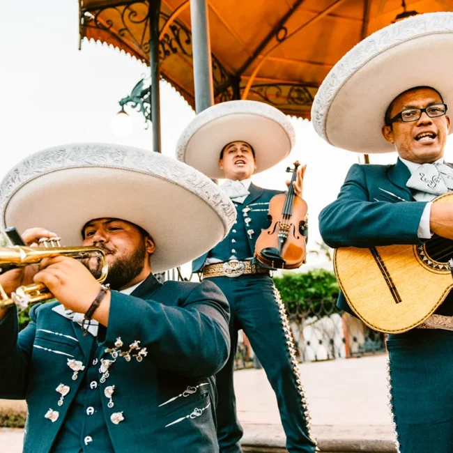 Drei Mariachi-Musiker tragen große weiße Sombreros und traditionelle Kleidung. Sie spielen Trompete, Geige und Gitarre.