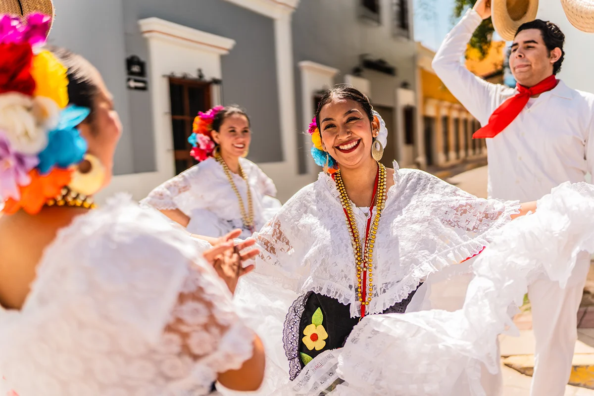 Drei mexikanische Tänzerinnen und ein Tänzer in traditioneller Kleidung tanzen zusammen. Die Frauen tragen Blumen in den Haaren, weiße Spitzenoberteile, Ketten und große Ohrringe.