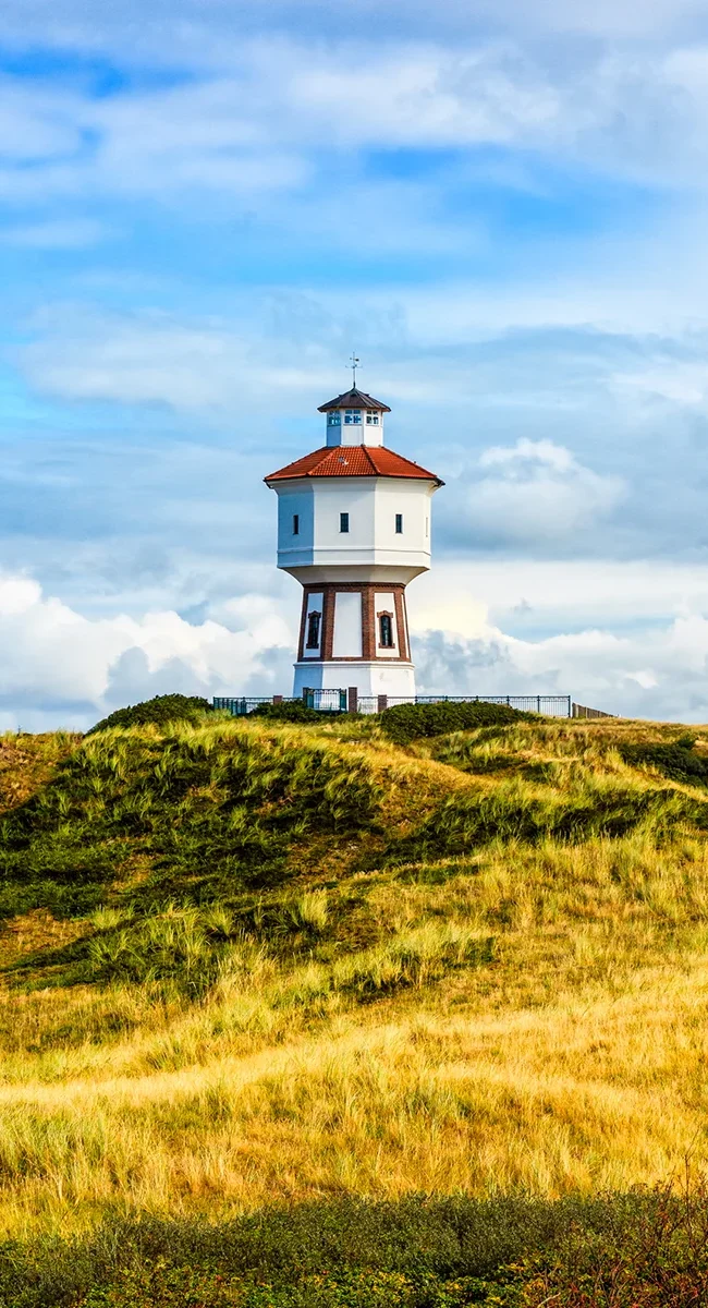 Der historische Wasserturm von Langeoog, das weiß-rote Wahrzeichen der Insel, wie er markant auf einer grasbewachsenen Düne unter einem leicht bewölkten blauen Himmel thront.