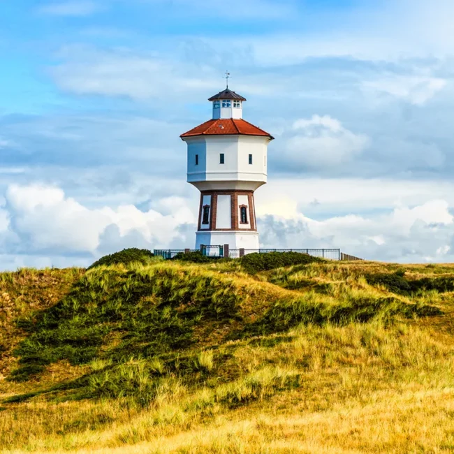 Der historische Wasserturm von Langeoog, das weiß-rote Wahrzeichen der Insel, wie er markant auf einer grasbewachsenen Düne unter einem leicht bewölkten blauen Himmel thront.