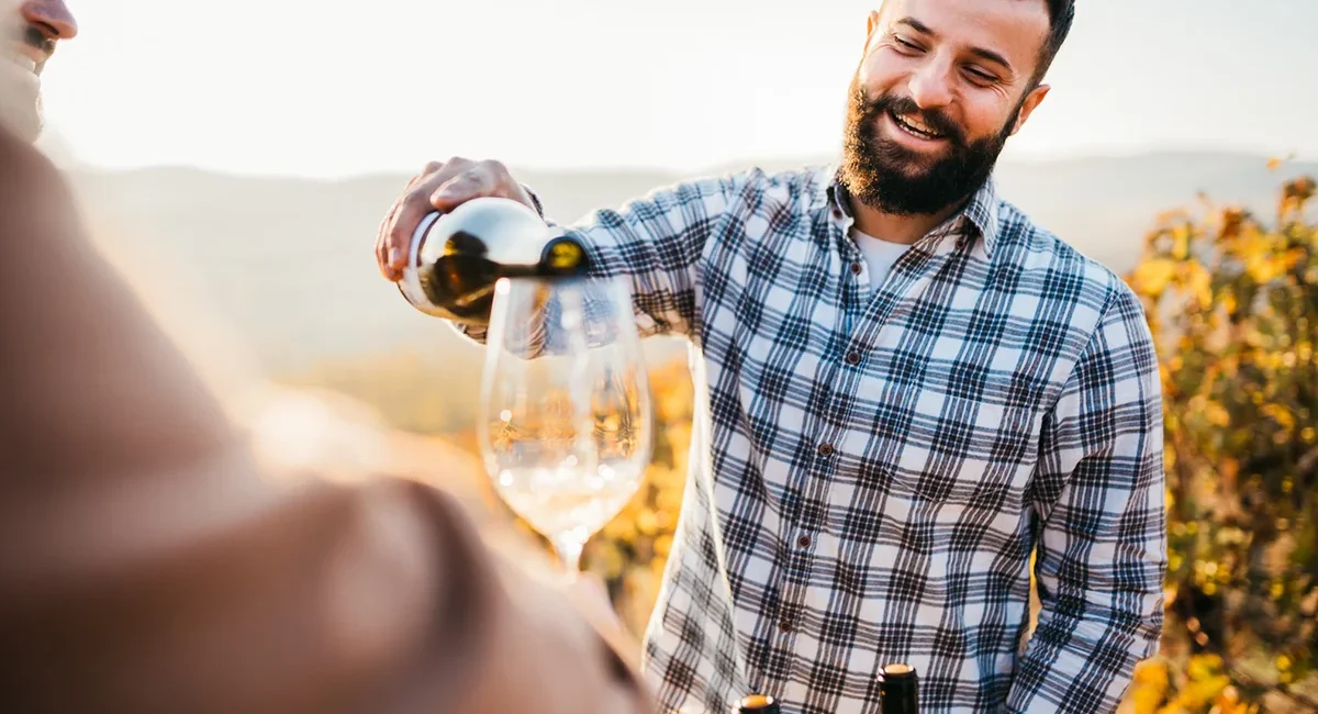 Winzer mit kariertem Hemd und Bart schenkt Wein bei einer Weinprobe im Weinberg ein