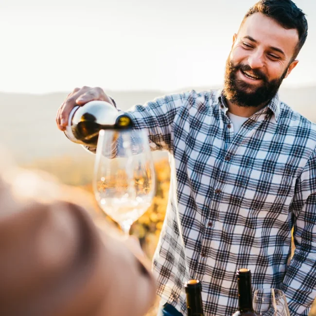 Winzer mit kariertem Hemd und Bart schenkt Wein bei einer Weinprobe im Weinberg ein
